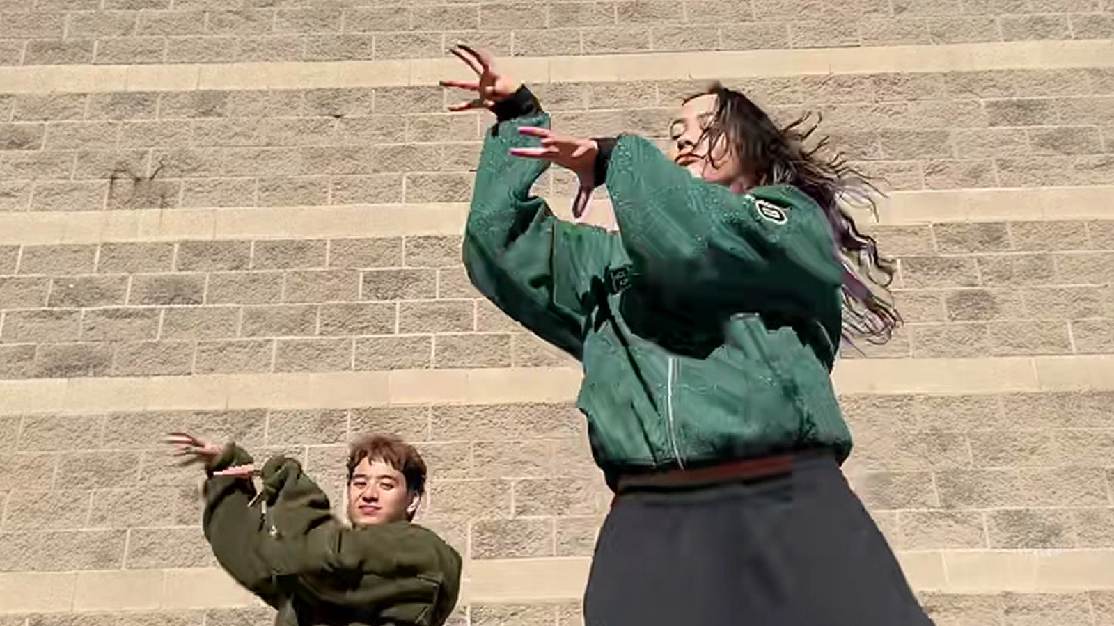 Two dancers in front of a sunny stone wall: the foreground lead on the right and the second dancer behind to the left.
