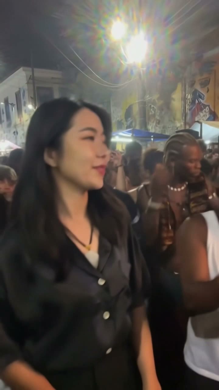 AI-generated woman in a black shirt dancing in the foreground of a street baile charme clip, with a man in a white tank top and brown hat behind her.