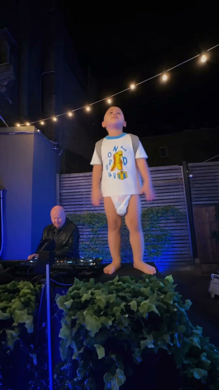 A child in a white dinosaur shirt standing on a lit stage in a night courtyard with string lights and a seated DJ.